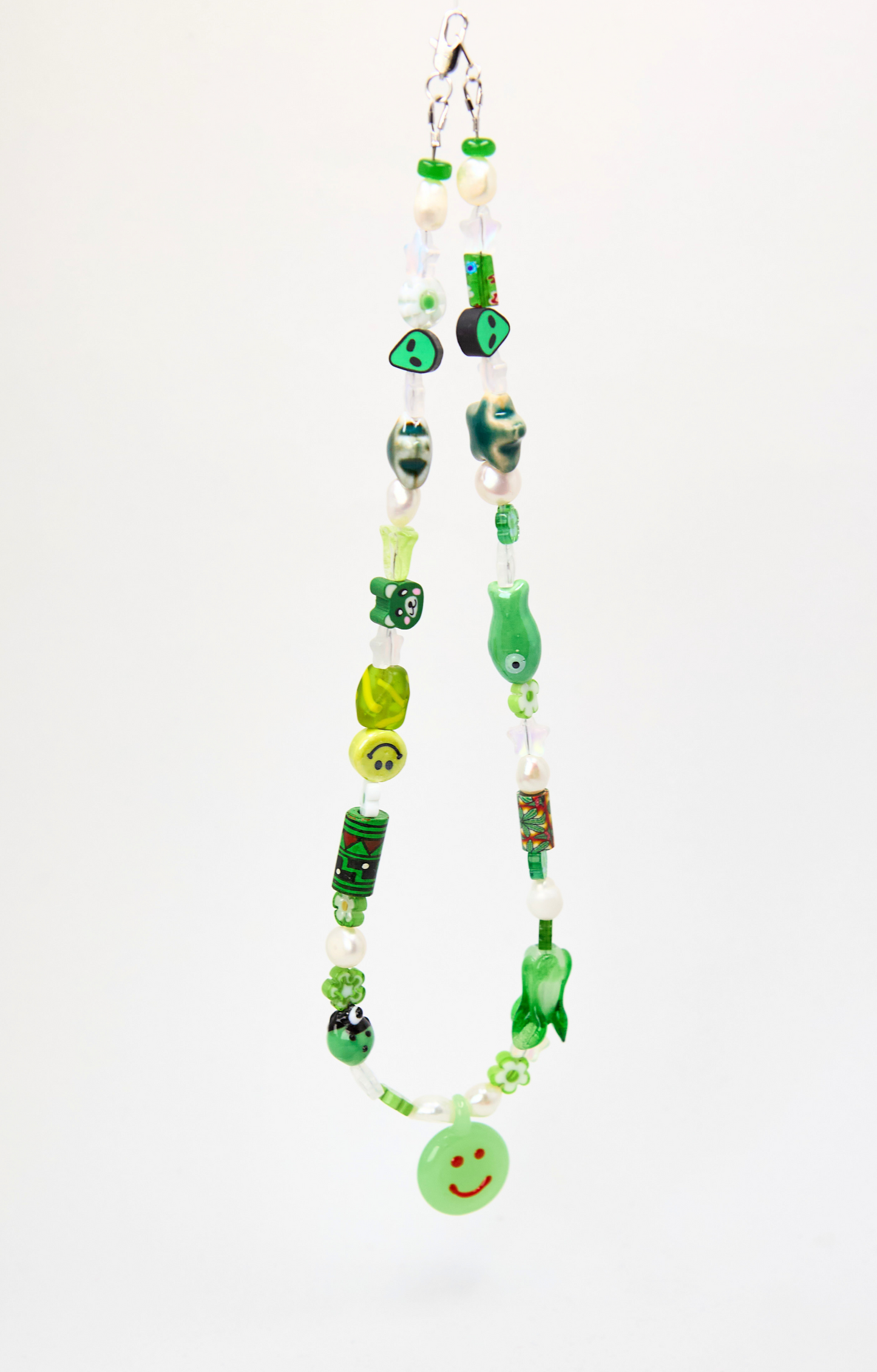 A handmade beaded necklace made from green glass, beads, and charms, featuring a small green smiley face button at the bottom, shown against a plain white background.