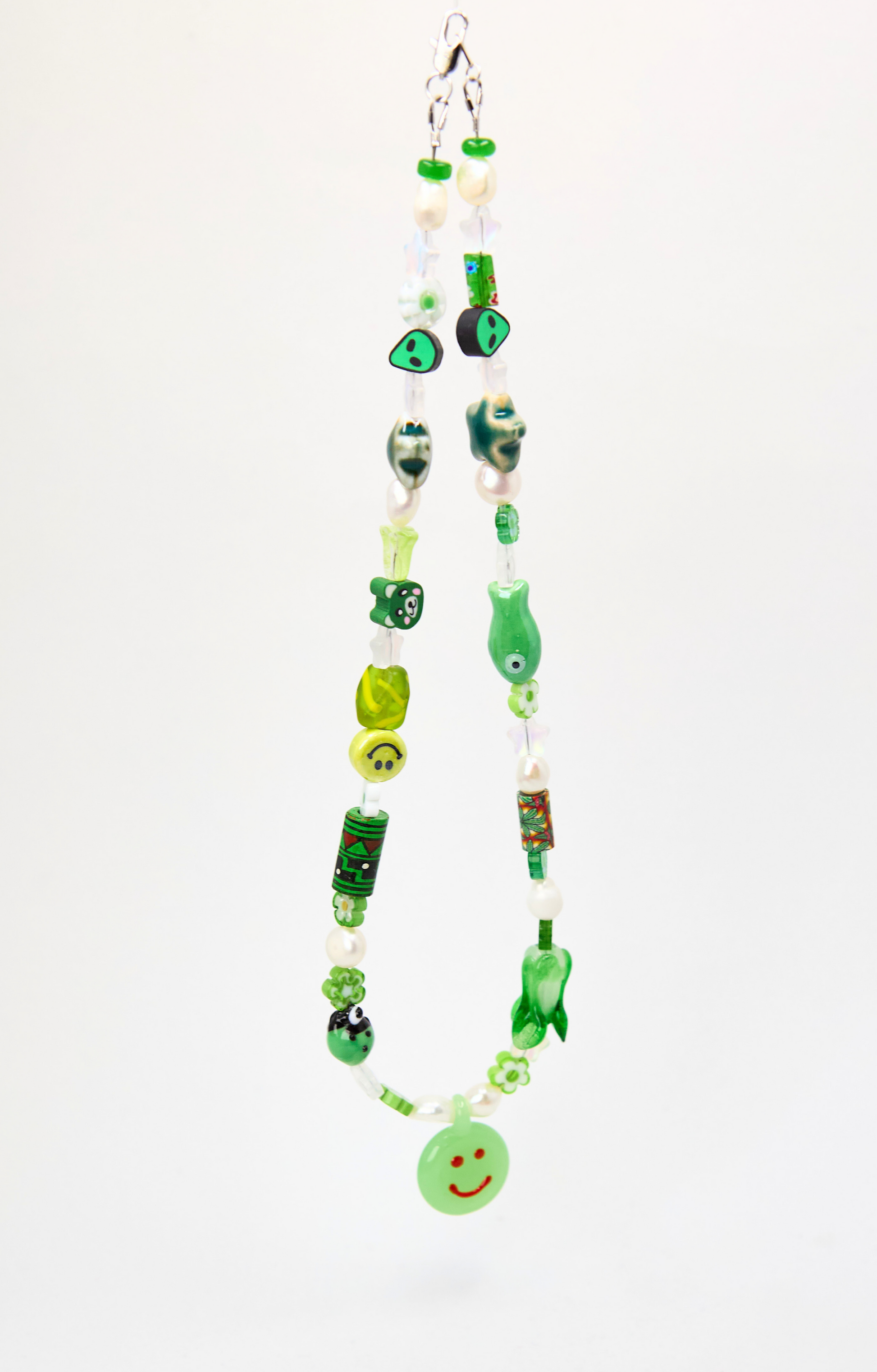 A handmade beaded necklace made from green glass, beads, and charms, featuring a small green smiley face button at the bottom, shown against a plain white background.