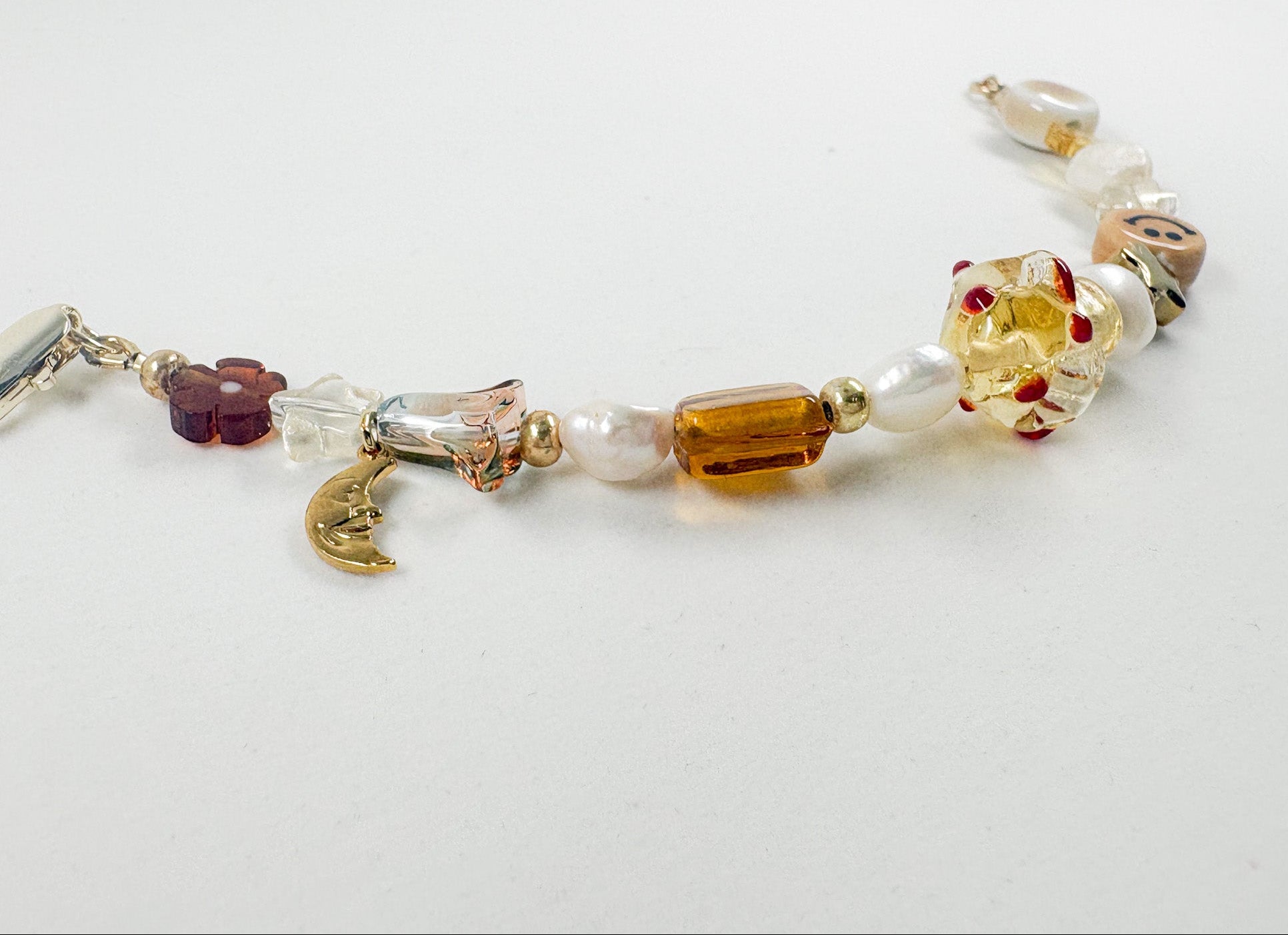 A handmade beaded bracelet featuring a mix of translucent amber, white pearl-like beads, a gold crescent moon charm, and a purple flower charm, displayed on a plain white surface.